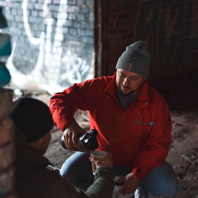 Ein Mann in einer roten Jacke der Johanniter und mit einer grauen Mütze gießt ein heißes Getränk aus einer Thermoskanne. Er hilft einem Obdachlosen.