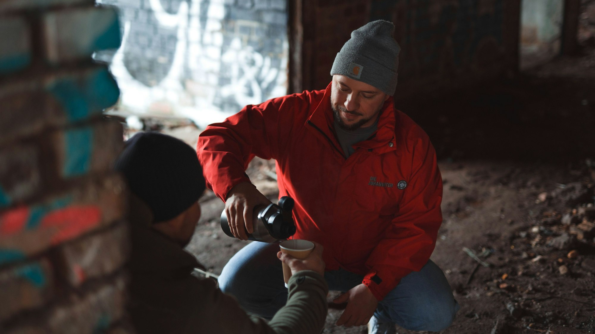 Ein Mann in einer roten Jacke der Johanniter und mit einer grauen Mütze gießt ein heißes Getränk aus einer Thermoskanne. Er hilft einem Obdachlosen.