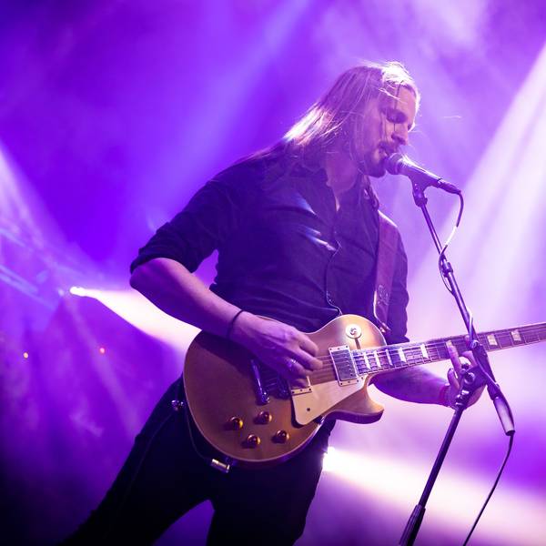 Laurent Teubl steht mit umgehängter Gitarre am Mikrofon und singt. Der Bühnenhintergrund ist in violettes Scheinwerferlicht getaucht.