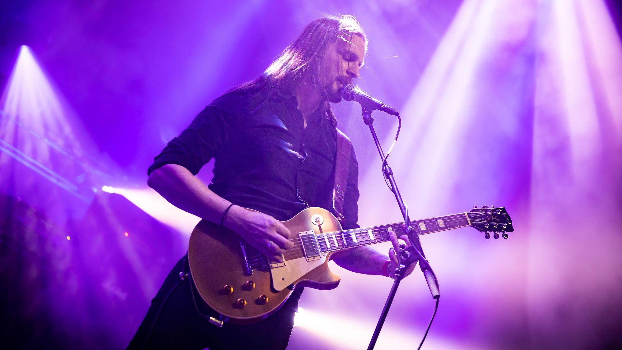 Laurent Teubl steht mit umgehängter Gitarre am Mikrofon und singt. Der Bühnenhintergrund ist in violettes Scheinwerferlicht getaucht.