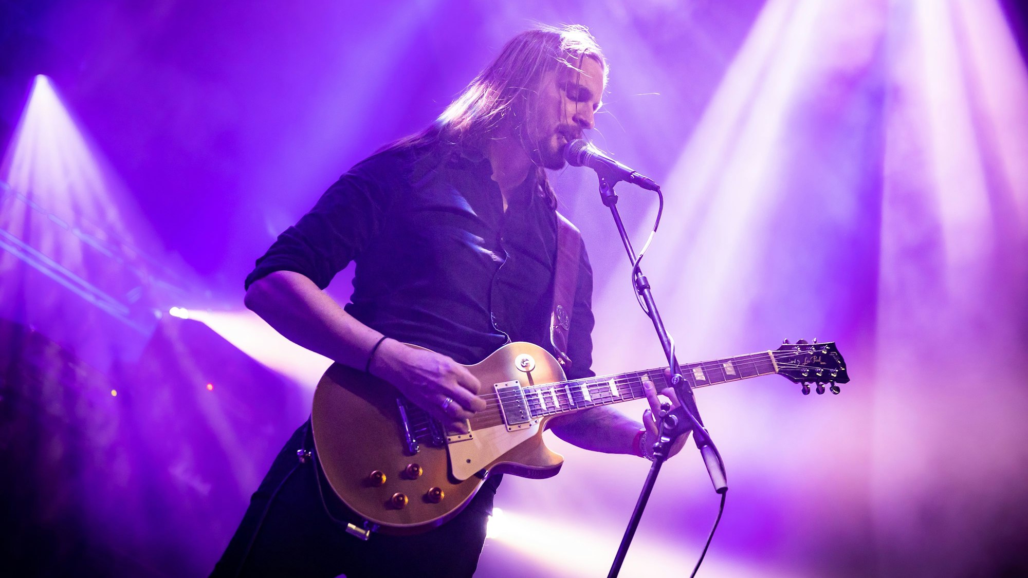 Laurent Teubl steht mit umgehängter Gitarre am Mikrofon und singt. Der Bühnenhintergrund ist in violettes Scheinwerferlicht getaucht.