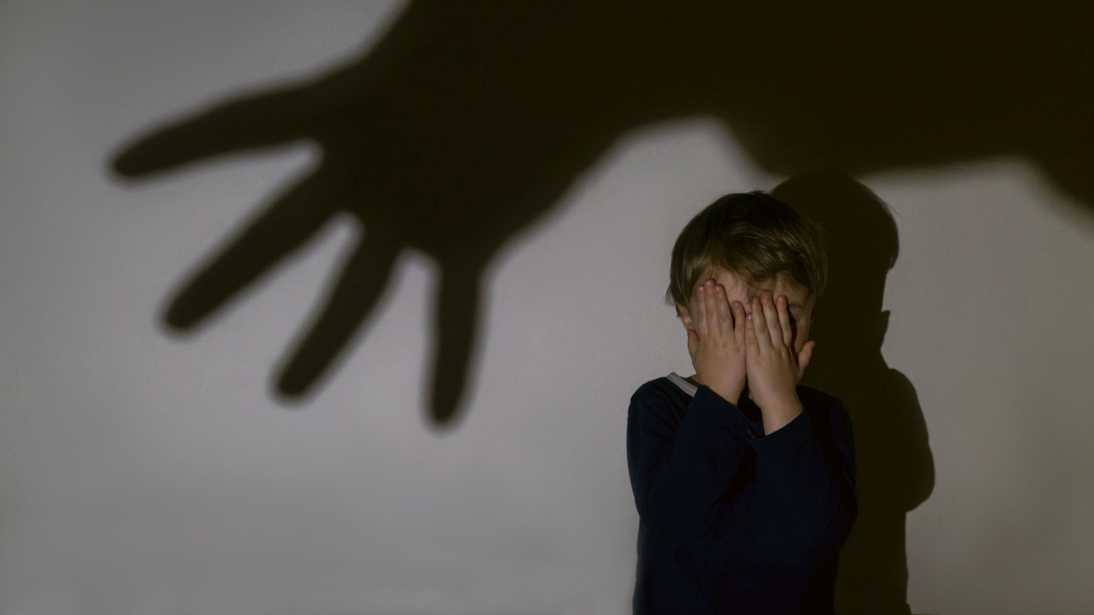Ein kleiner Junge sitzt vor einer Wand und hält vor Schrecken seine Hände vors Gesicht. An der Wand ist ein großer Schatten einer ausgestreckten Hand zu sehen.