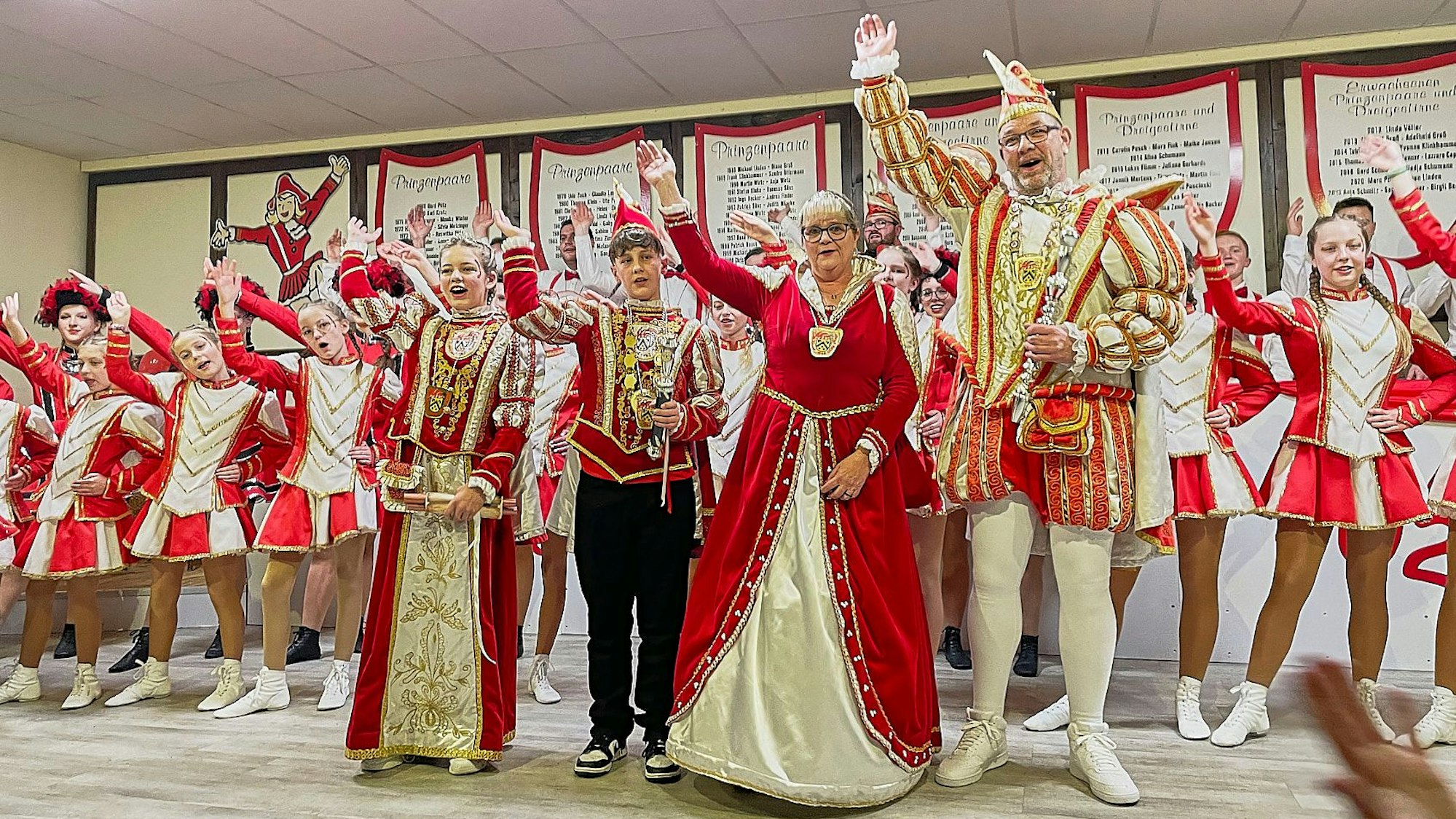 Auf der Bühne stehen die beiden Prinzenpaare, dahinter zahlreiche Gardetänzerinnen und -tänzer, alle in rot-weißen Kostümen.
