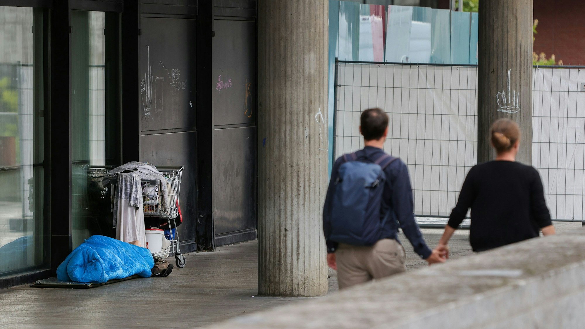 Obdachloser liegt in einem Schlafsack vor der Fassade am Römisch-Germanischen Museum. Ein Paar geht Händchen haltend daran vorbei.