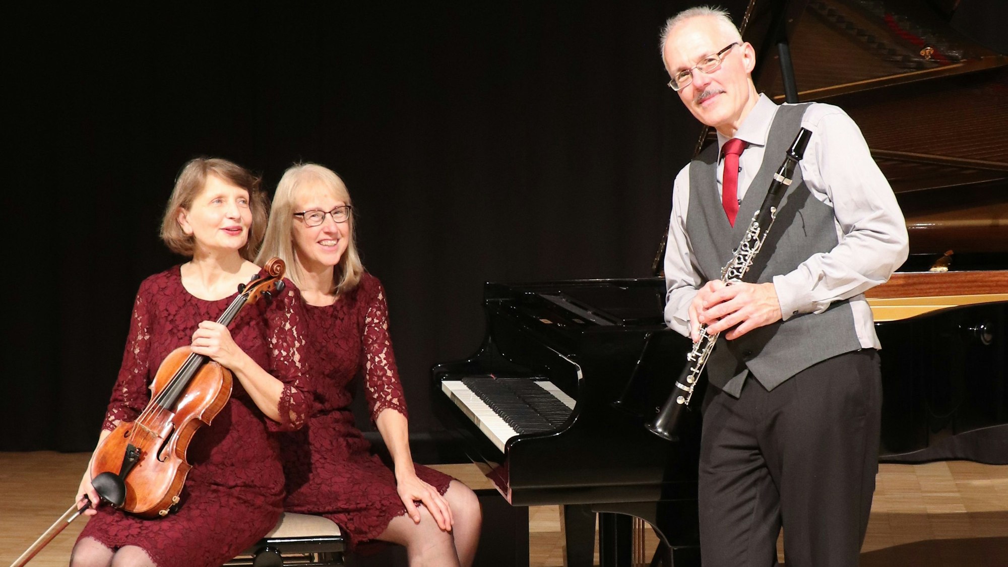 Zwei Frauen und ein Mann sind um einen Flügel versammelt.-