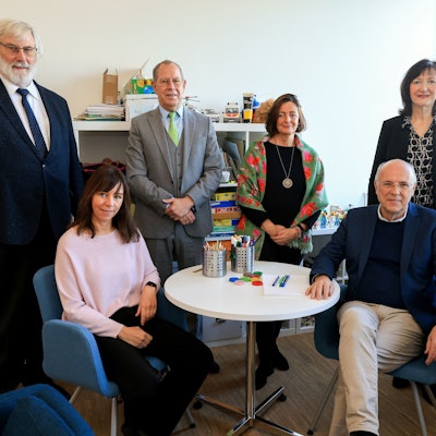 Das Bild zeigt drei Frauen und drei Männer in schickerer Kleidung, die an einem kleinen runden Tisch sitzen oder stehen und in die Kamera schauen