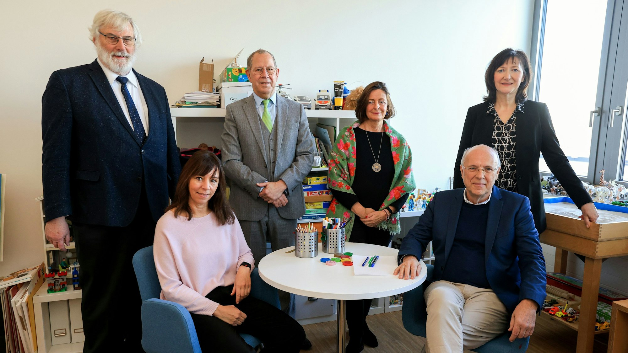 Das Bild zeigt drei Frauen und drei Männer in schickerer Kleidung, die an einem kleinen runden Tisch sitzen oder stehen und in die Kamera schauen