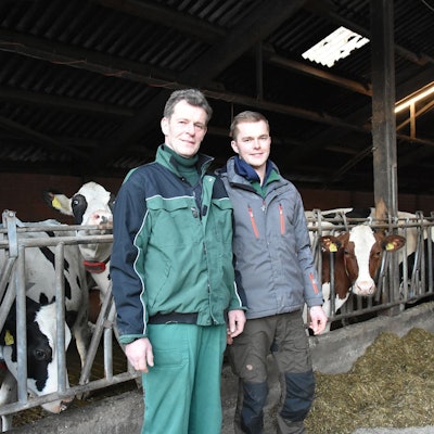Ein Mann und sein Sohn stehen in einem Stall. Im Hintergrund sind mehrere Kühe zu sehen.