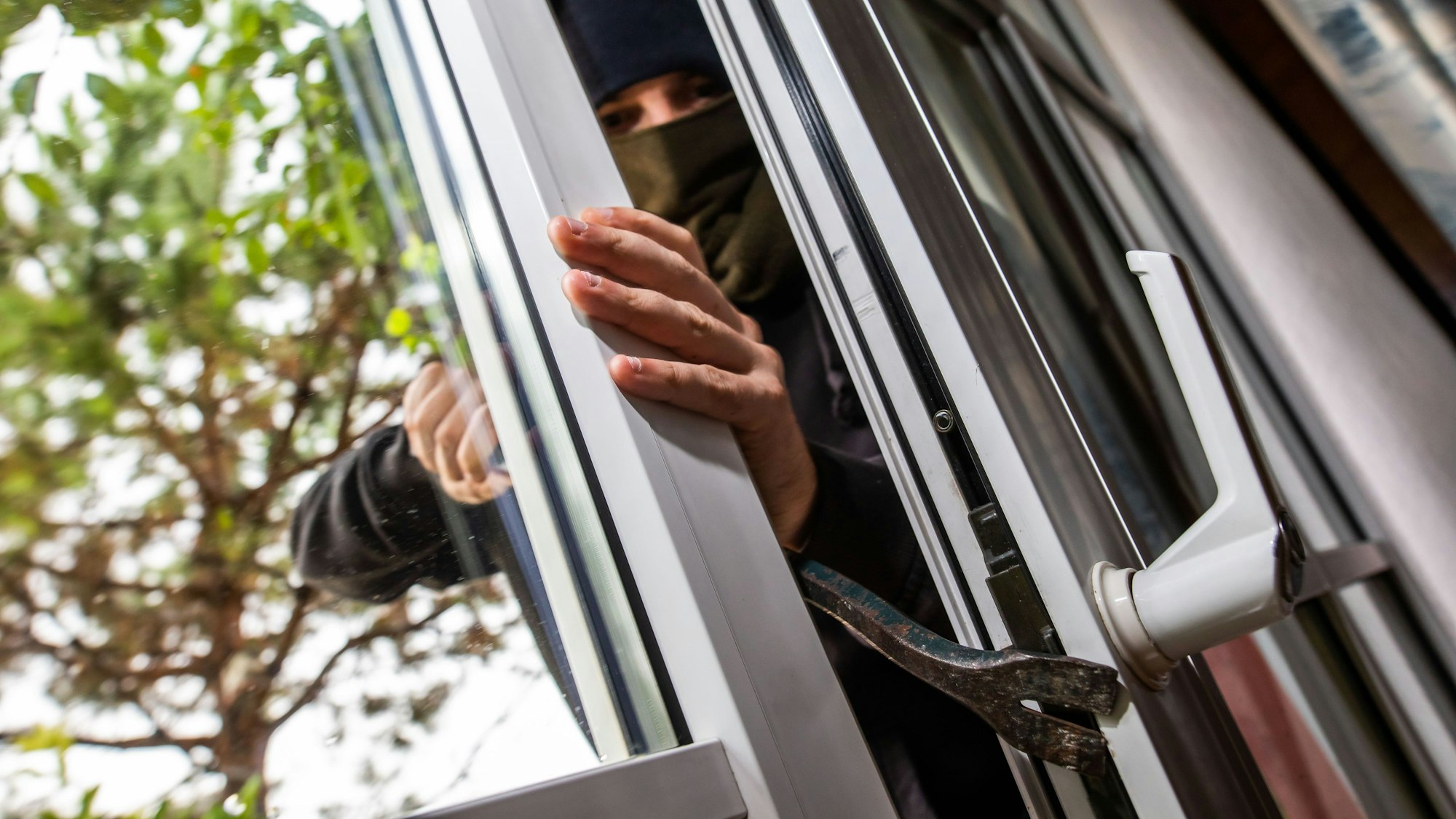 Ein Mann mimt einen Einbrecher und benutzt ein Brecheisen, um eine gekippte Terrassentür aufzuhebeln.