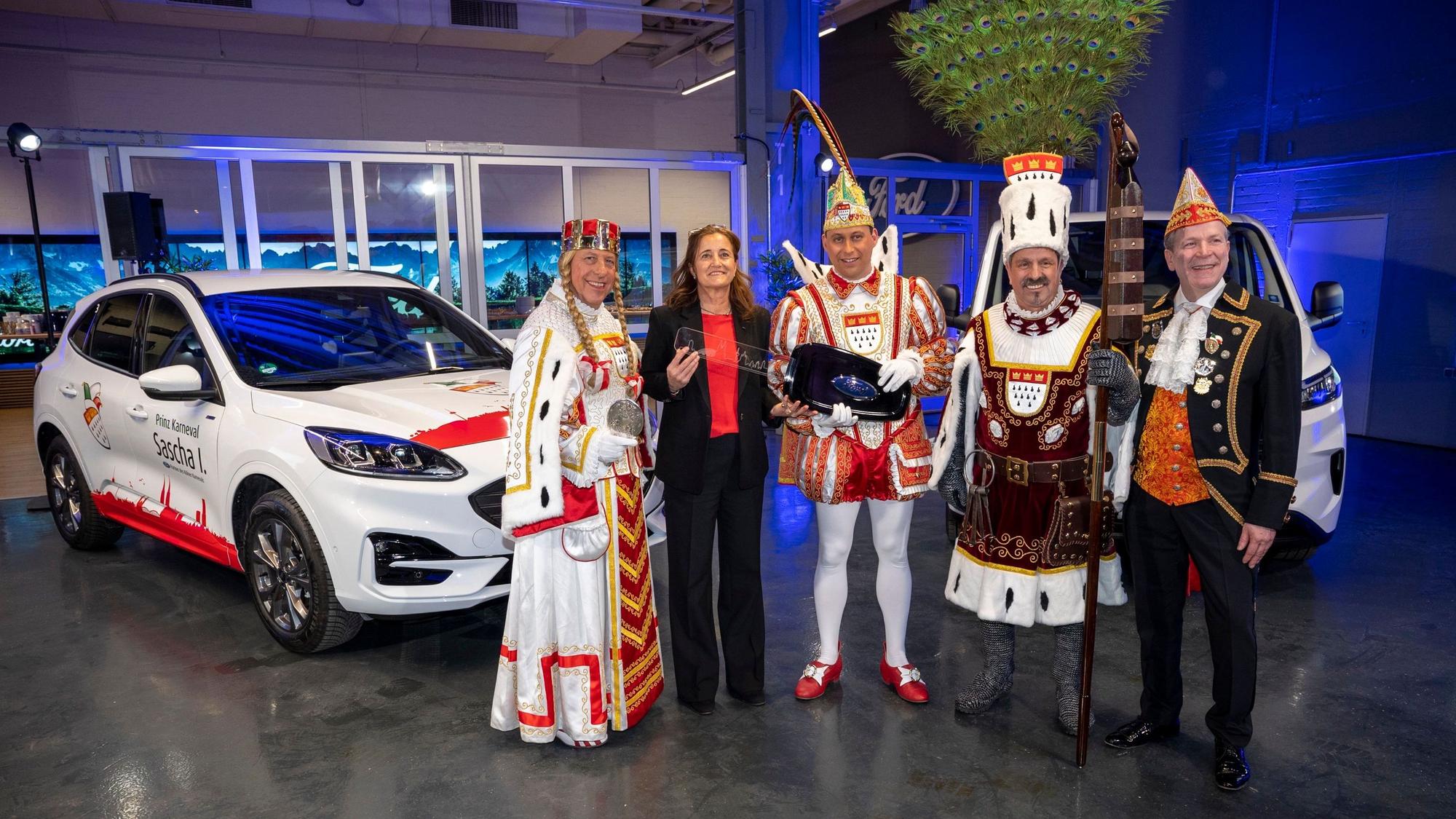 Das Dreigestirn im Ornat, Claudia Vogt und Christoph Kuckelkorn stehen vor einem weißen Ford Kuga.