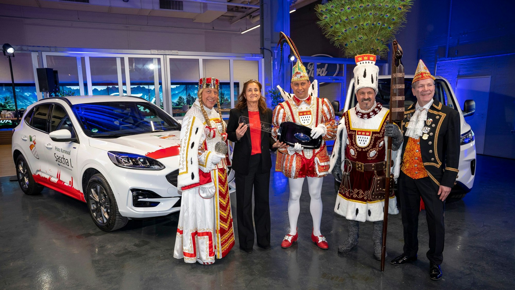 Das Dreigestirn im Ornat, Claudia Vogt und Christoph Kuckelkorn stehen vor einem weißen Ford Kuga.