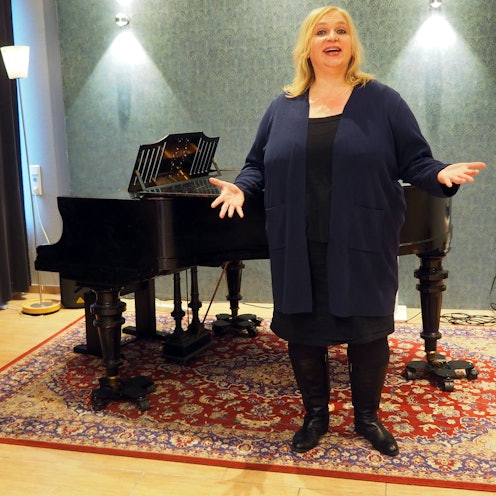 Eine blonde Frau steht mit ausgebreiteten Armen vor einem Flügel.