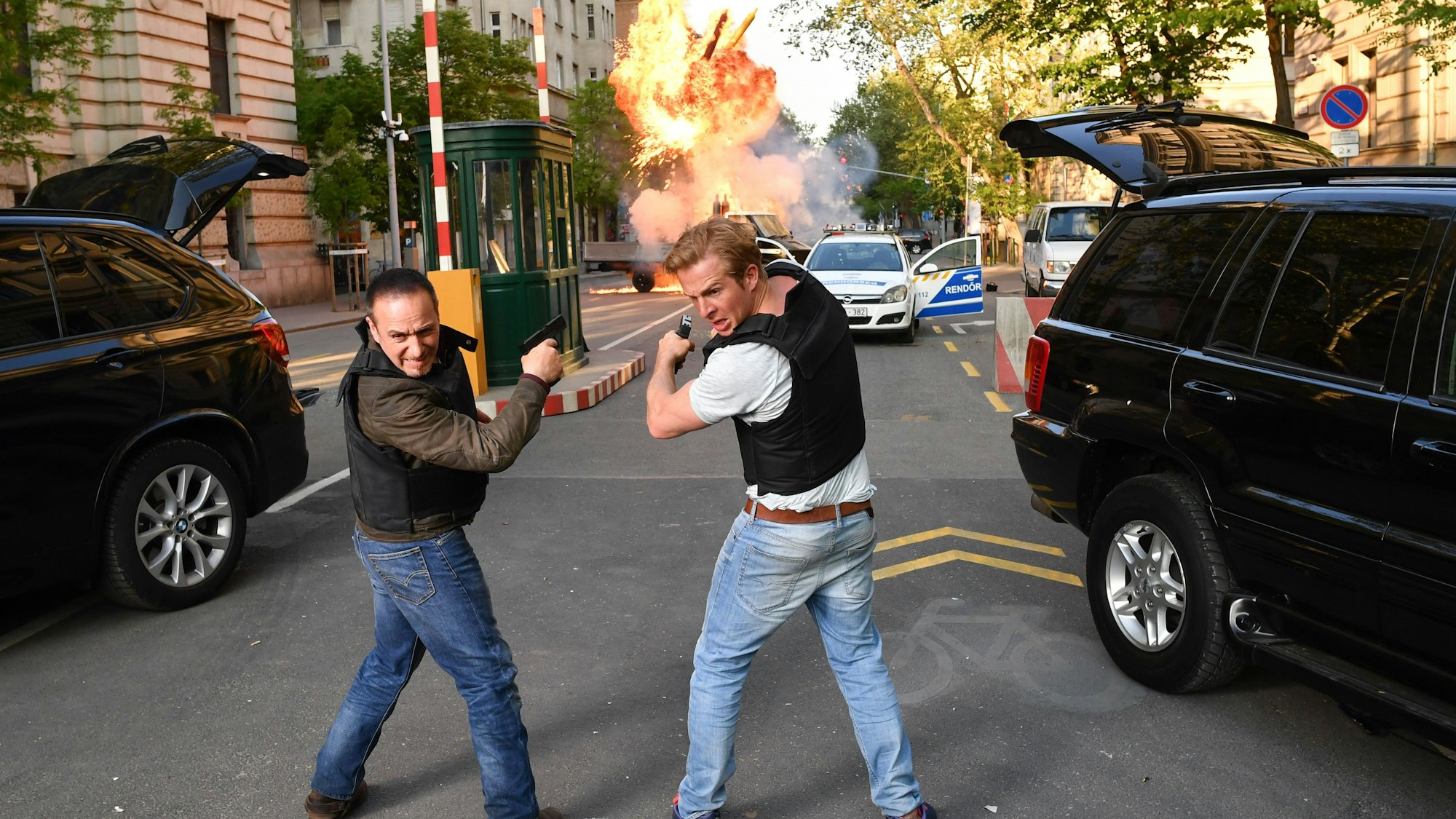 Die Schauspieler Erdogan Atalay (l) als Semir Gerkhan und Daniel Roesner als Paul Renner stehen im Zentrum der ungarischen Hauptstadt bei einer inszenierten Explosion für die RTL-Action-Serie „Alarm für Cobra 11 - Die Autobahnpolizei“ im Set der Folge „Most Wanted“. Rechts und links im Bild stehen zwei schwarze Limousinen mit geöffneten Kofferräumen. Mittig hinten im Bild steht ein Polizeiauto, um das herum eine Explosion stattfindet.