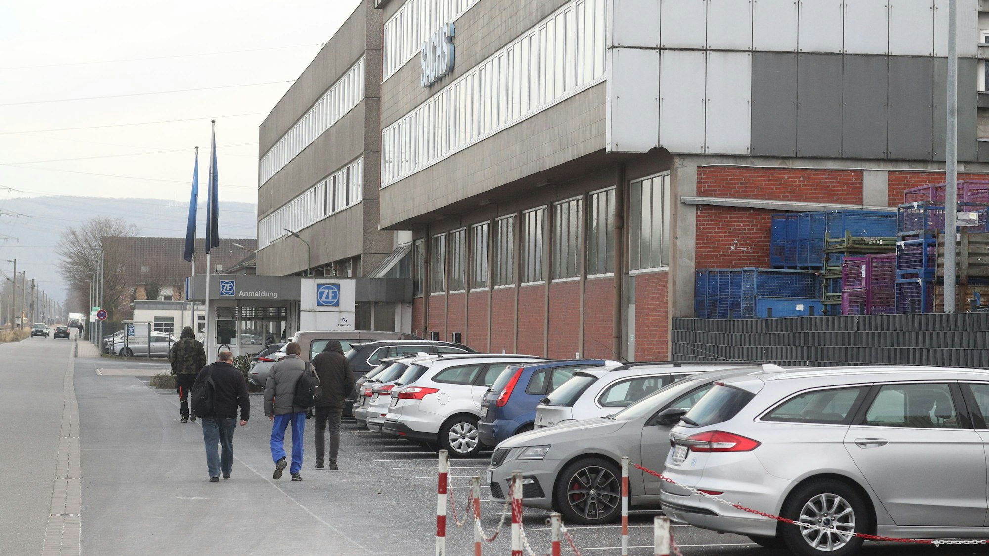 Menschen in blauer Arbeitskleidung laufen an einer Industriehalle entlang, das Logo von ZF ist auf einer Tafel am angebracht.