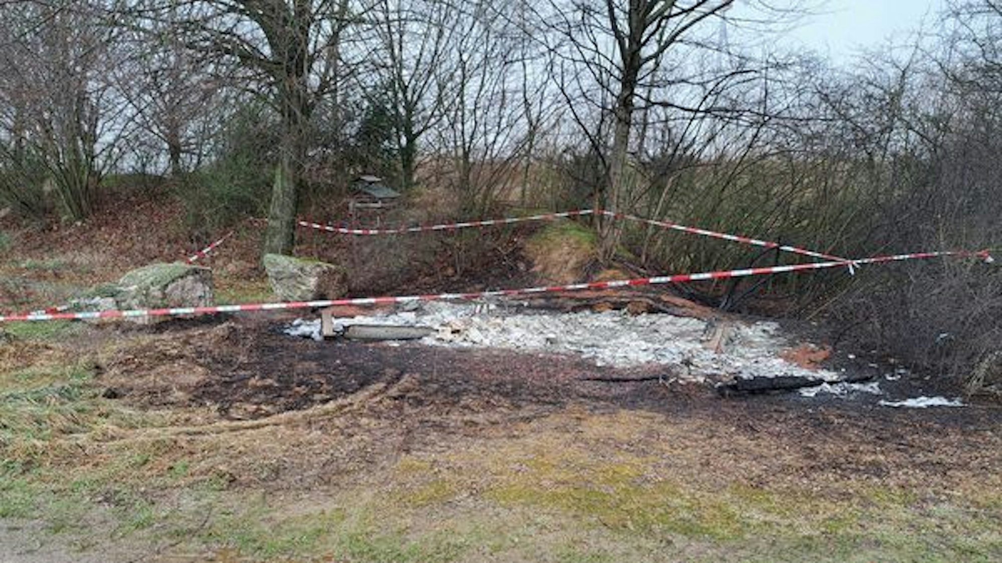 Auf dem Foto ist die abgebrannte Hütte am Gedenkplatz zu sehen. Der Bereich ist mit rot-weißem Flatterband abgesperrt.