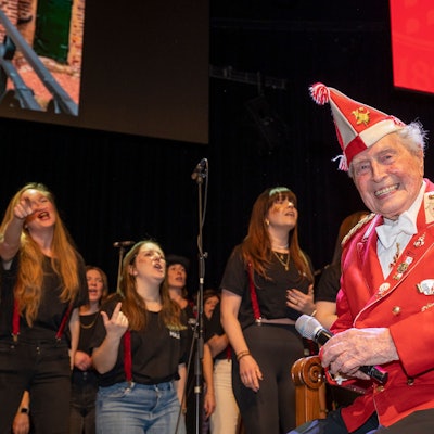 Ludwig Sebus sitzt auf einem großen Stuhl auf der Bühne und hört den Sängerinnen zu.