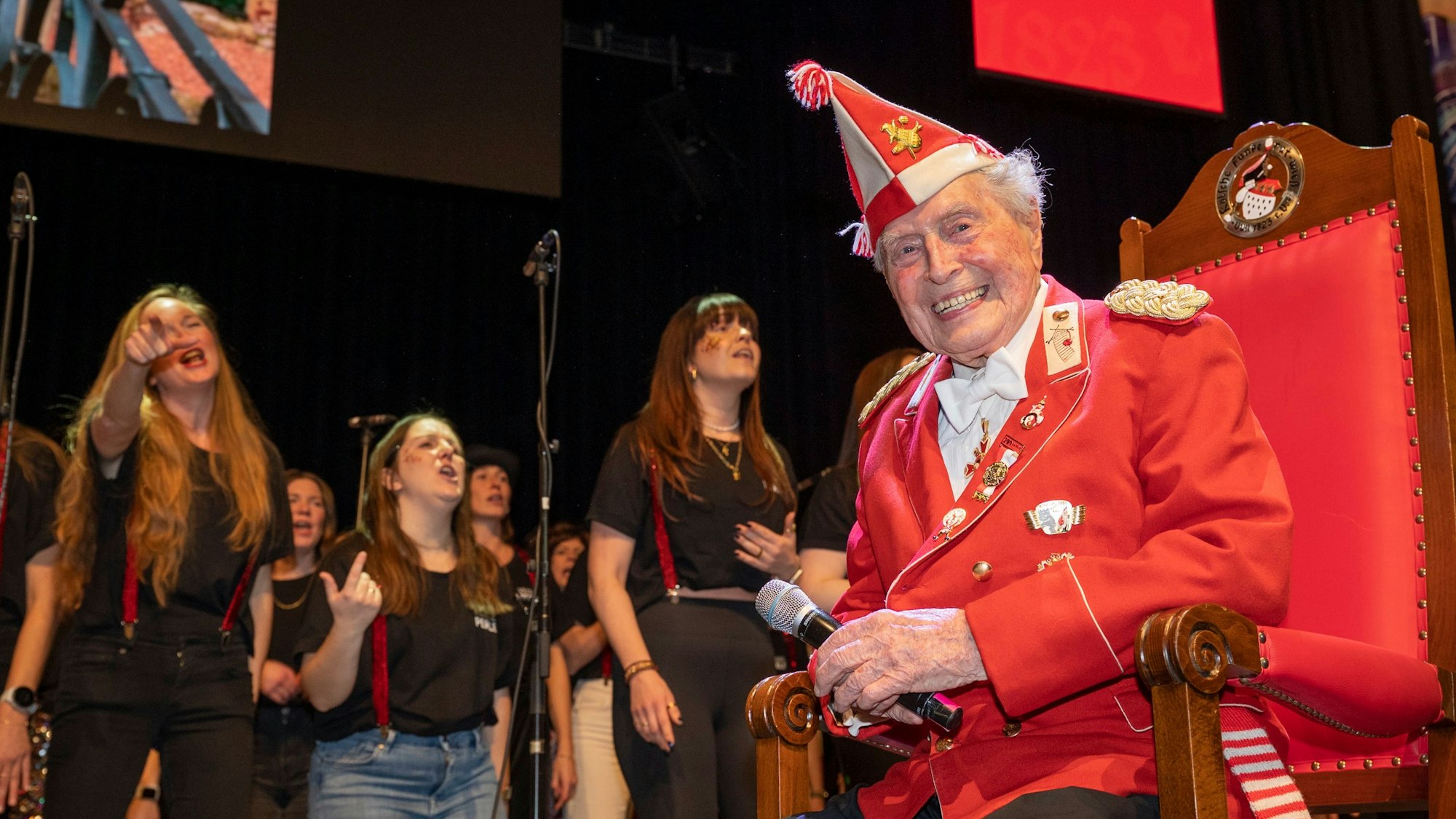 Ludwig Sebus sitzt auf einem großen Stuhl auf der Bühne und hört den Sängerinnen zu.