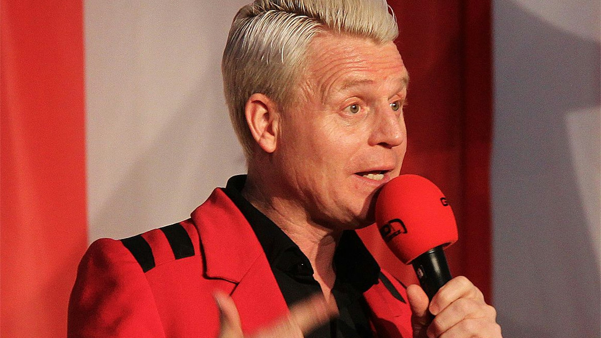 Guido Cantz trägt einen roten Anzug und hält ein rotes Mikrofon in der Hand.
