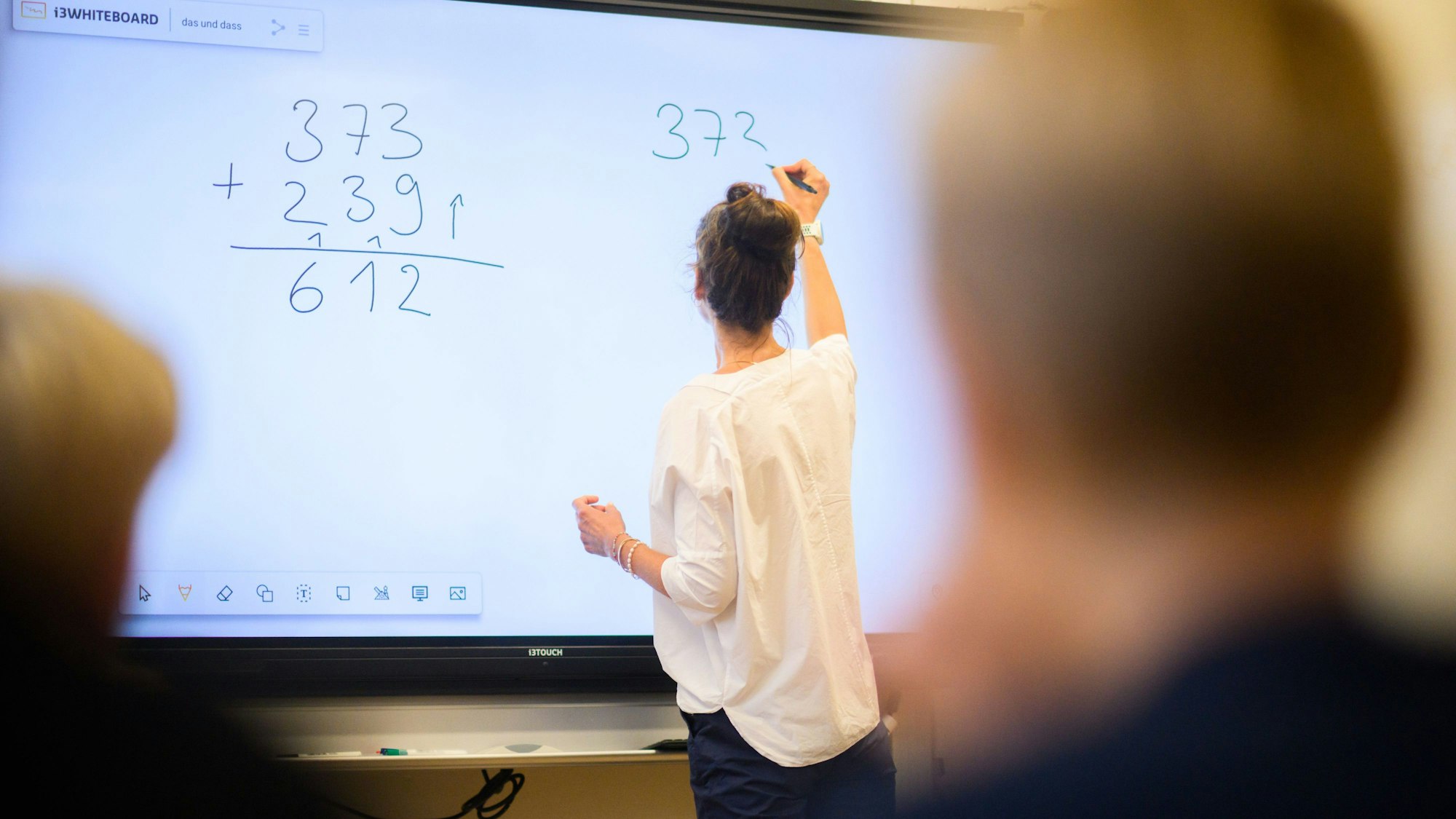 Eine Lehrerin schreibt eine Mathematikaufgabe auf eine digitale Schultafel im Klassenraum einer 4. Klasse einer Grundschule.