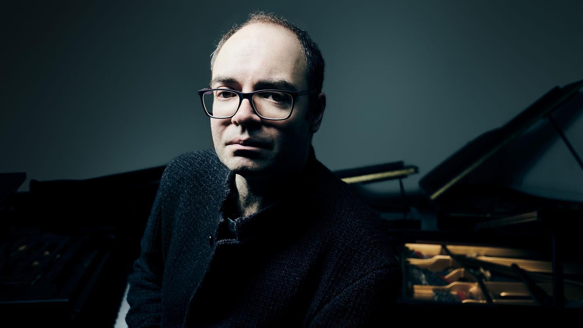 Alexander Melnikov trägt eine eckige Brille und schaut ernst in die Kamera. Sein Gesicht ist hell erleuchtet, der Hintergrund, wo auch ein Flügel steht, ist dunkel.