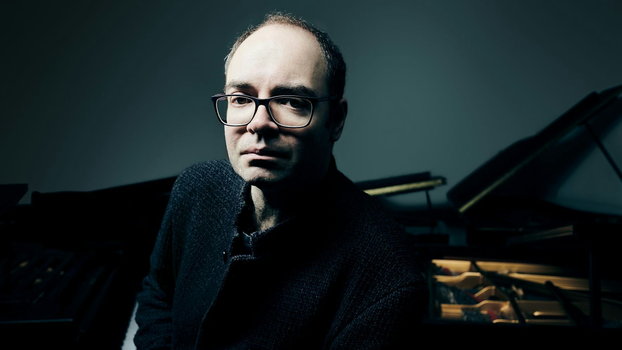 Alexander Melnikov trägt eine eckige Brille und schaut ernst in die Kamera. Sein Gesicht ist hell erleuchtet, der Hintergrund, wo auch ein Flügel steht, ist dunkel.