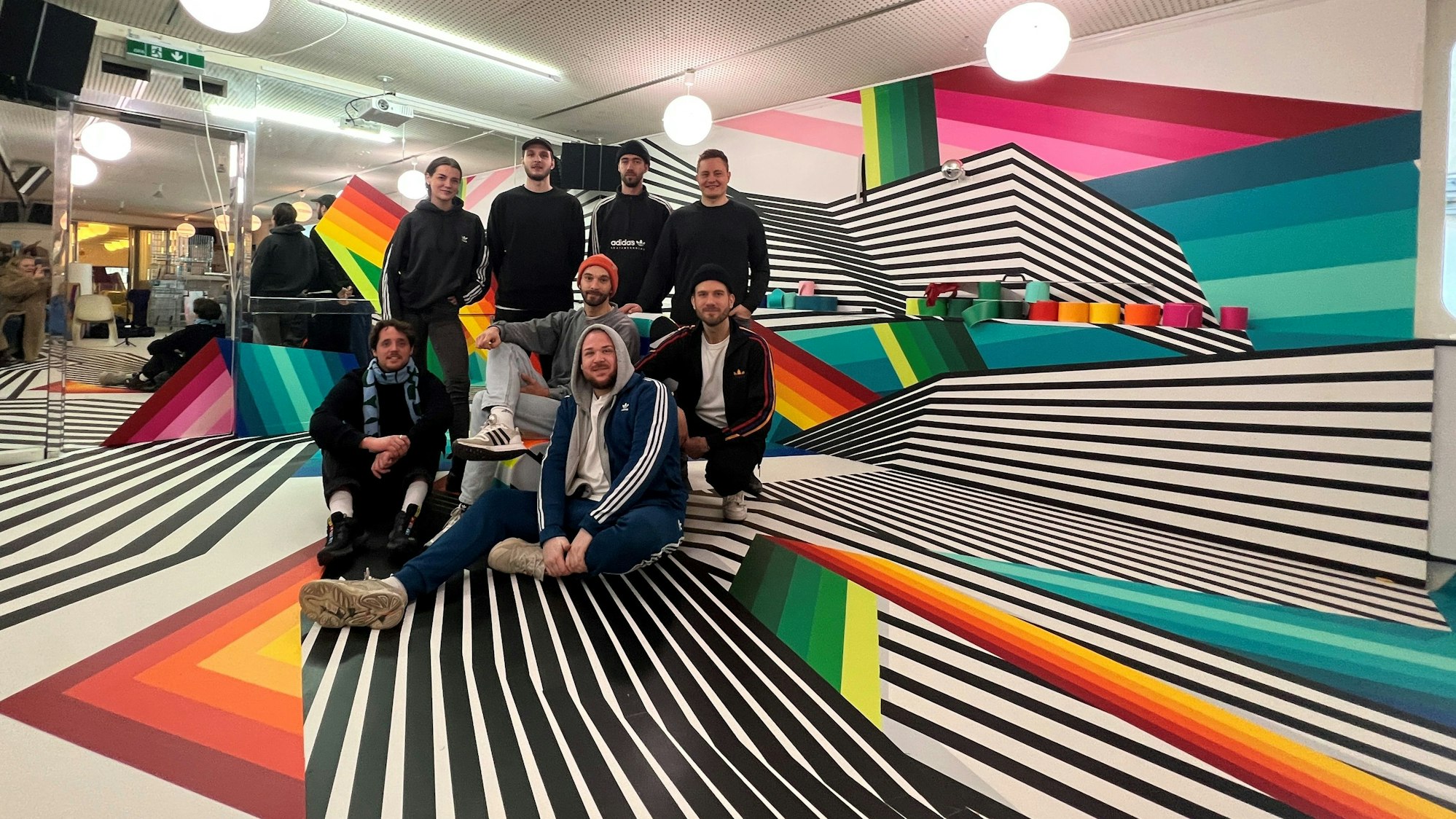 Eine Gruppe junger Männder in schwarzen Kapuzenjacken sitzt und steht in einem futuristisch gestalteten Interieur.