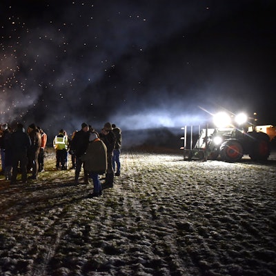 Eine Gruppe Menschen steht im Dunkeln an einem großen Feuer, rechts im Bild ist ein Traktor mit angeschaltetem Licht zu sehen.