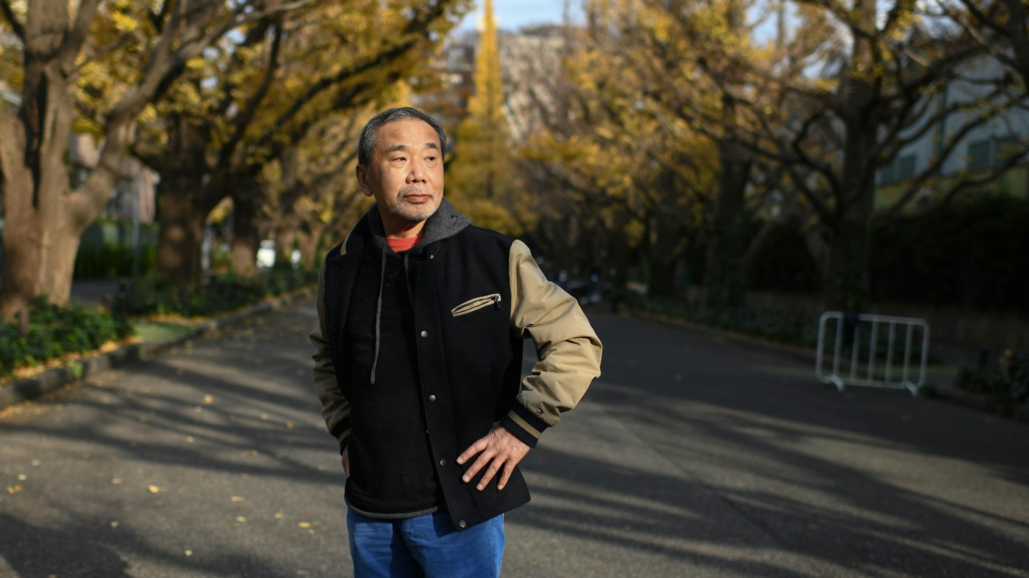 Porträt des Autors Haruki Murakami, der eine Collegejacke trägt und in einer Allee in Tokyo steht.