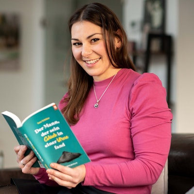 Das Bild zeigt Sara Schütze auf ihrer Couch. In den Händen hält sie ihren eigenen Roman.