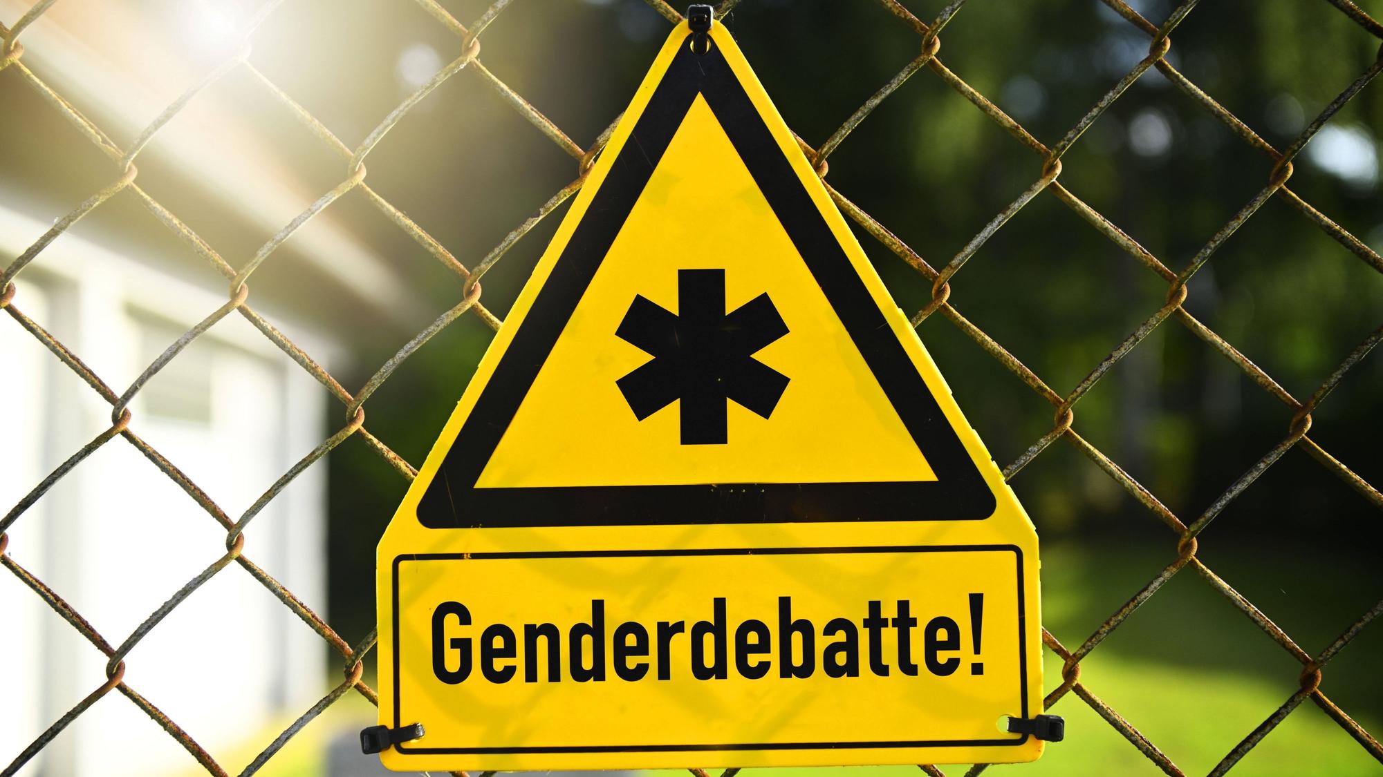 Ein gelbes Gefahrenschild hängt an einem Drahtzaun. Es ist mit einem Gendersternchen in schwarz umrandetem Dreieck beschriftet und der Aufschrift „Genderdebatte“ unterschrieben. Es handelt sich hierbei um eine Fotomontage.