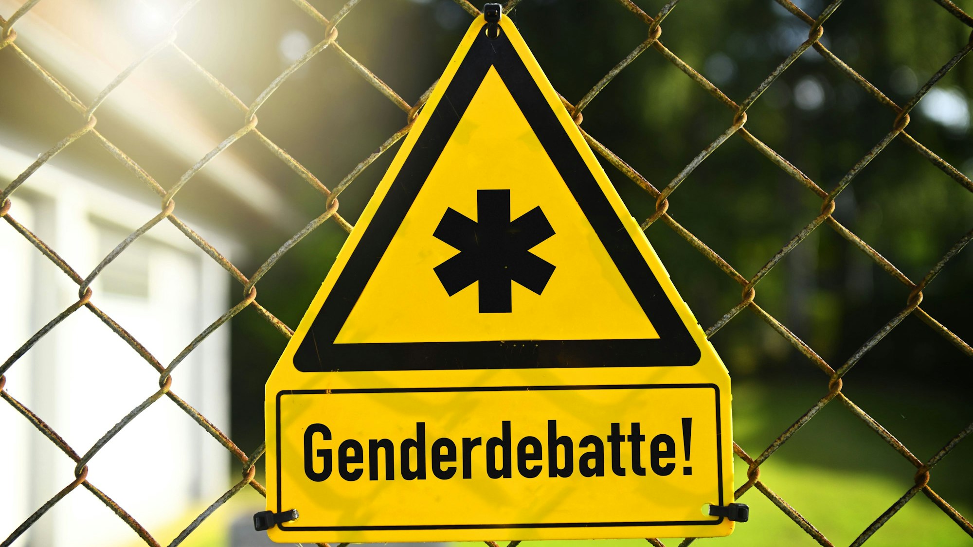 Ein gelbes Gefahrenschild hängt an einem Drahtzaun. Es ist mit einem Gendersternchen in schwarz umrandetem Dreieck beschriftet und der Aufschrift „Genderdebatte“ unterschrieben. Es handelt sich hierbei um eine Fotomontage.