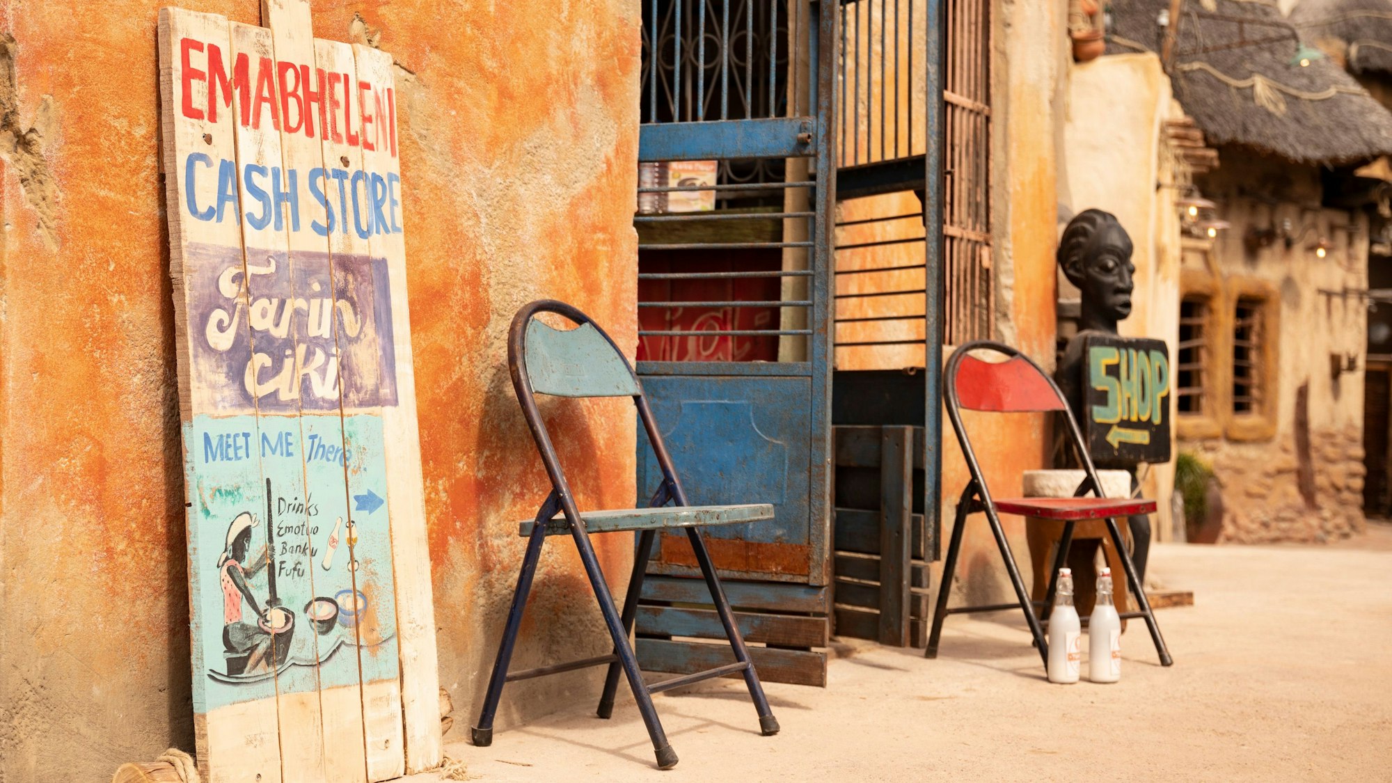 Vor einem rustikalen Haus mit Lehmwänden stehen neben einer offenen Türe zwei staubige Stühle. Im Hintergrund sind weitere traditionelle Lehmhäuser zu sehen.