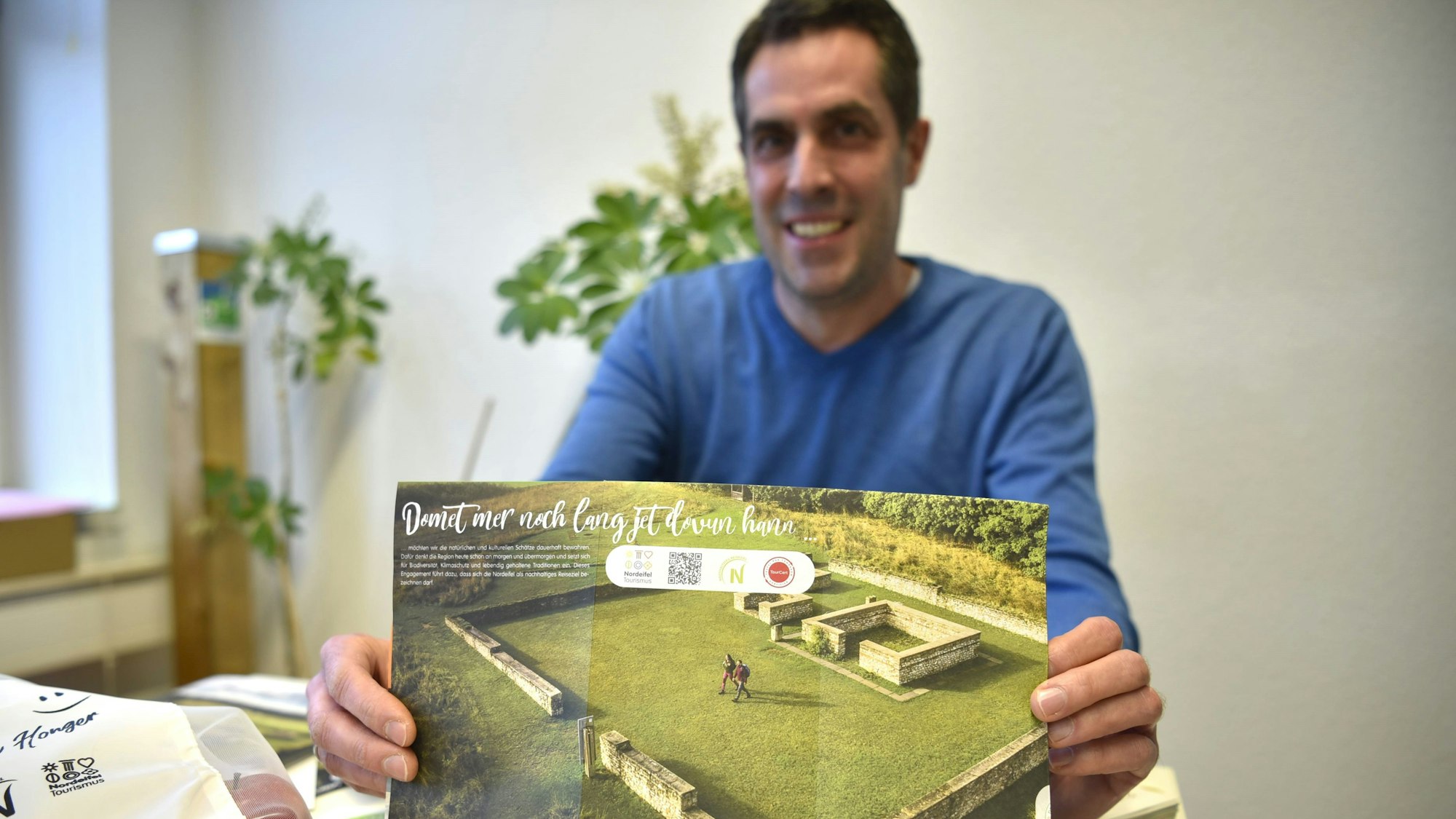 Patrick Schmidder, Geschäftsführer der Nordeifel Tourismus GmbH, hält einen Flyer in die Kamera, auf dem ein Luftbild zusammen mit dem Schriftzug „Domet mer noch lang jet dovun hann“ abgebildet ist.