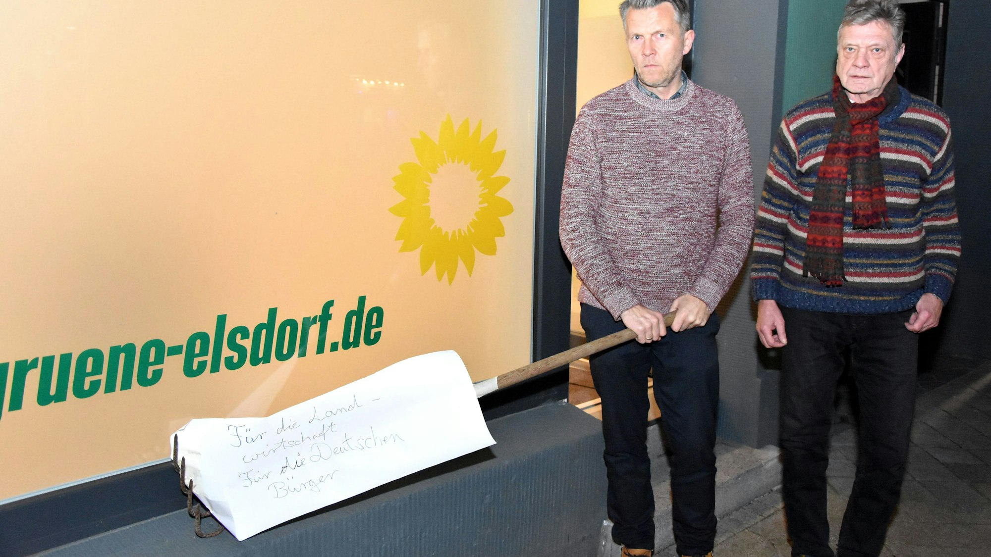 Auf dem Foto sind die Elsdorfer Grünen-Politiker Michael Broich und Hartmut Bergheim vor dem Büro ihrer Fraktion zu sehen. Broich hält die Harke in den Händen.