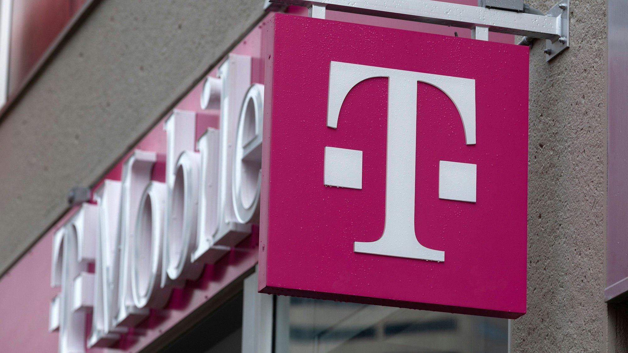 Ein Logo der Deutschen Telekom bzw. T-Mobile ist vor einem Geschäft in einer Innenstadt zu sehen.