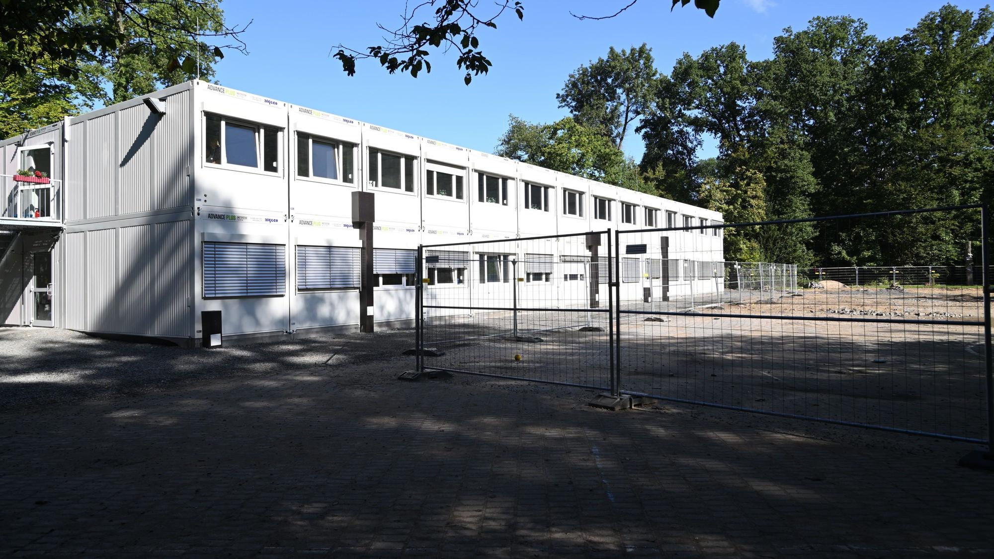 Ein doppelstöckiger, grauer Containerbau auf einem Schulhof. Davor ein Bauzaun.