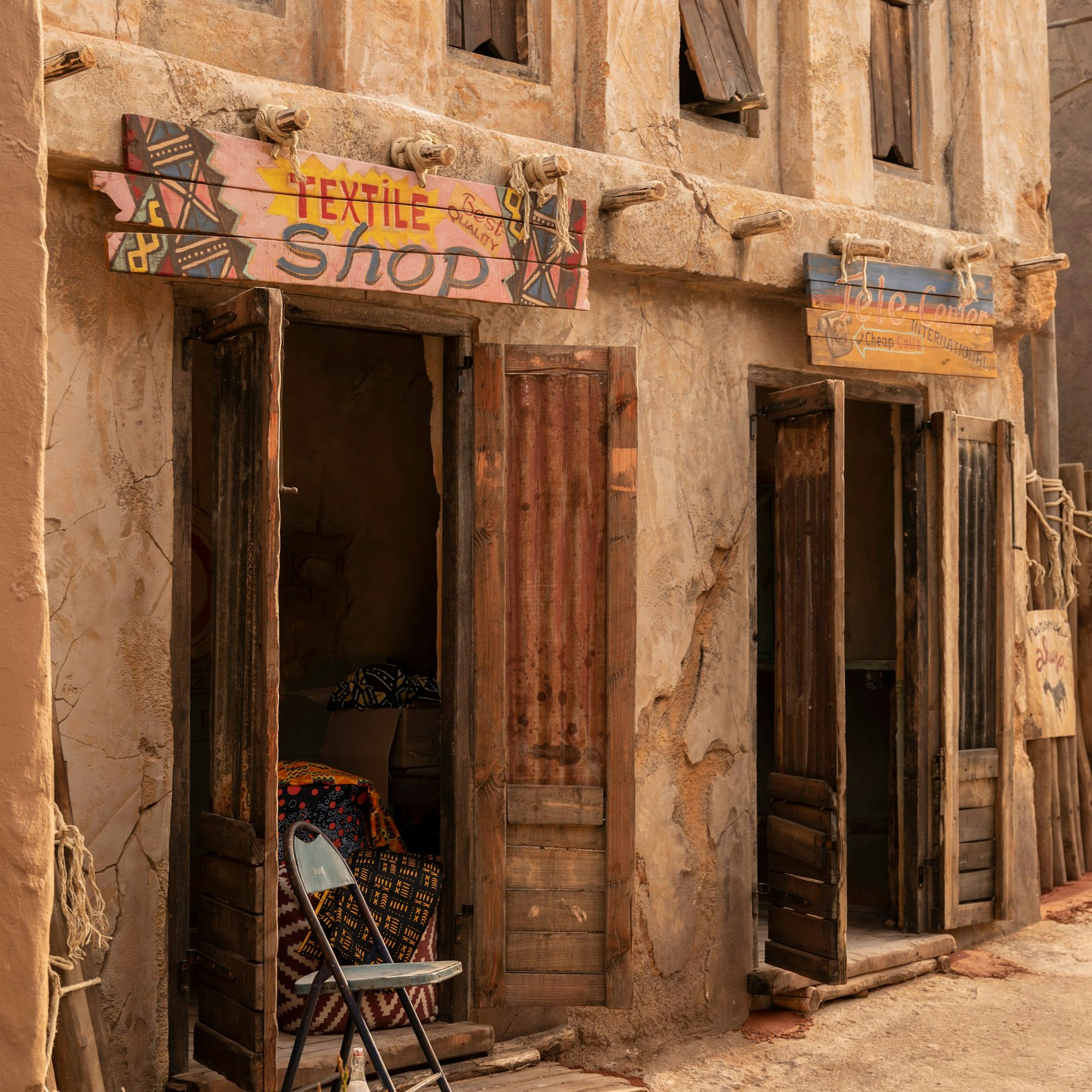 Vor einem sandigen Weg sind Lehmhütten mit Ladenschildern zu sehen. Ihre altertümlich anmutenden Holztüren stehen weit offen.