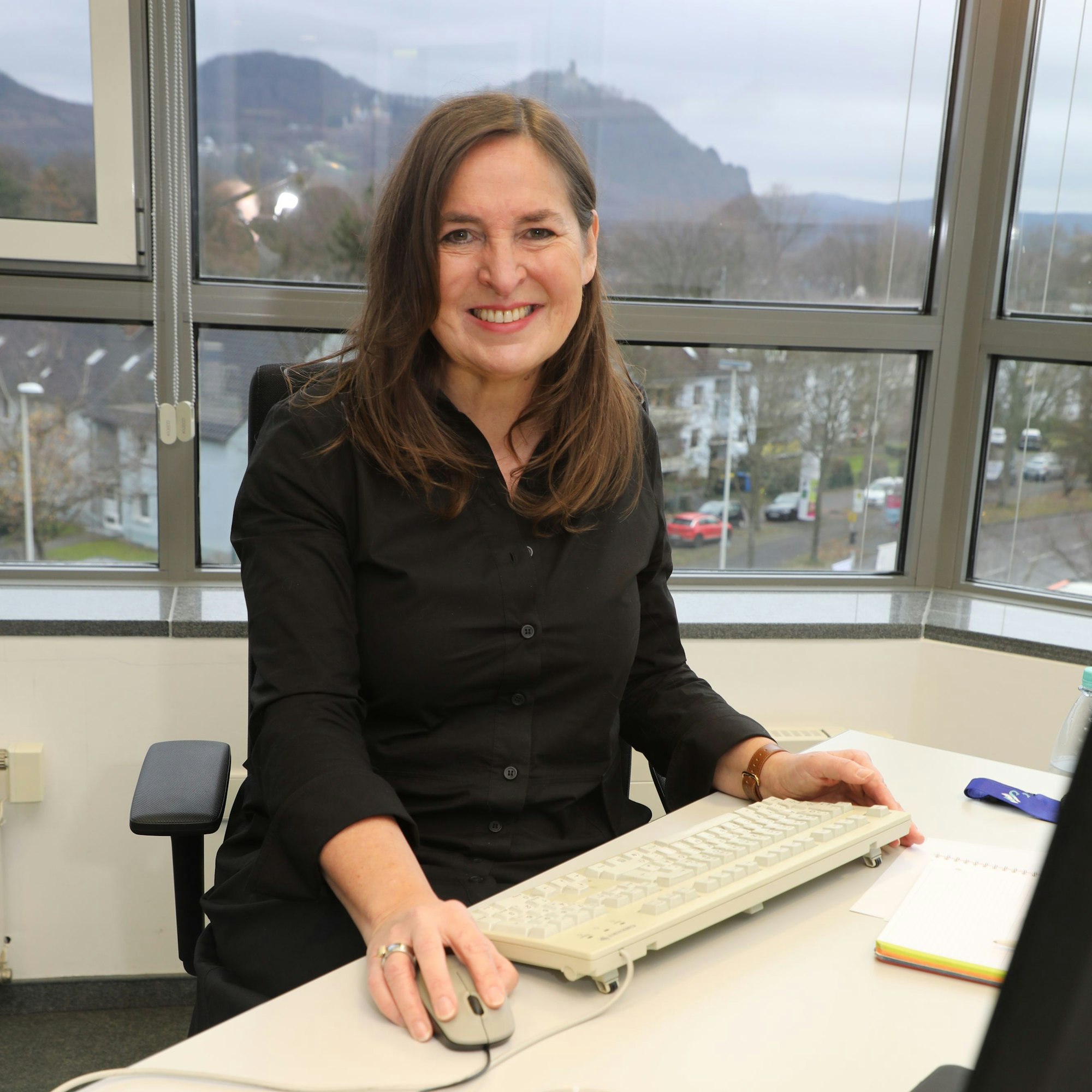 Jungfrau Sabine sitzt an einem Schreibtisch, im Hintergrund ist der Drachenfels zu sehen.