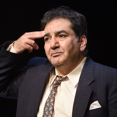 Szene aus dem Stück „Ich werde nicht hassen“, der Schauspieler Mohammad-Ali Behboudi trägt einen dunkelblauen Anzug und Krawatte. Er hält sich zwei Finger wie einen Pistolenlauf an die Schläfe.