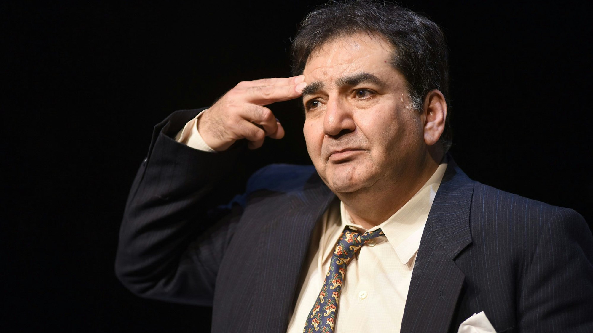 Szene aus dem Stück „Ich werde nicht hassen“, der Schauspieler Mohammad-Ali Behboudi trägt einen dunkelblauen Anzug und Krawatte. Er hält sich zwei Finger wie einen Pistolenlauf an die Schläfe.