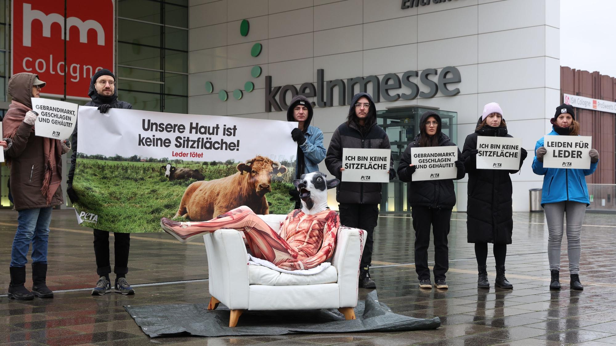 Demonstrierende von Peta mit Plakaten vor dem Eingang der Deutzer Messe. Auf einem weißen Ledersofa sitzt eine Person in einem Morphsuit, der aussieht wie ein gehäuteter Körper, mit Rindermaske.