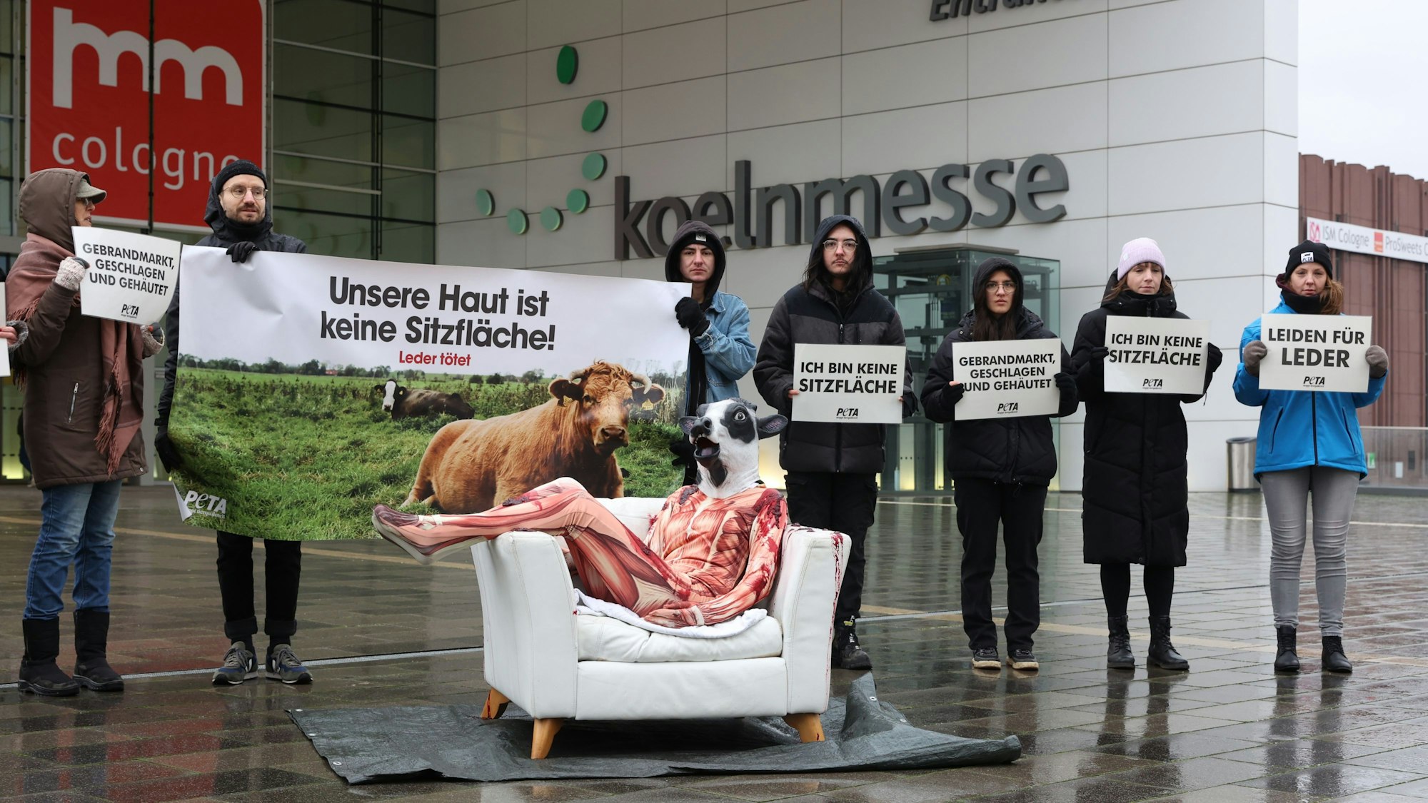 Demonstrierende von Peta mit Plakaten vor dem Eingang der Deutzer Messe. Auf einem weißen Ledersofa sitzt eine Person in einem Morphsuit, der aussieht wie ein gehäuteter Körper, mit Rindermaske.