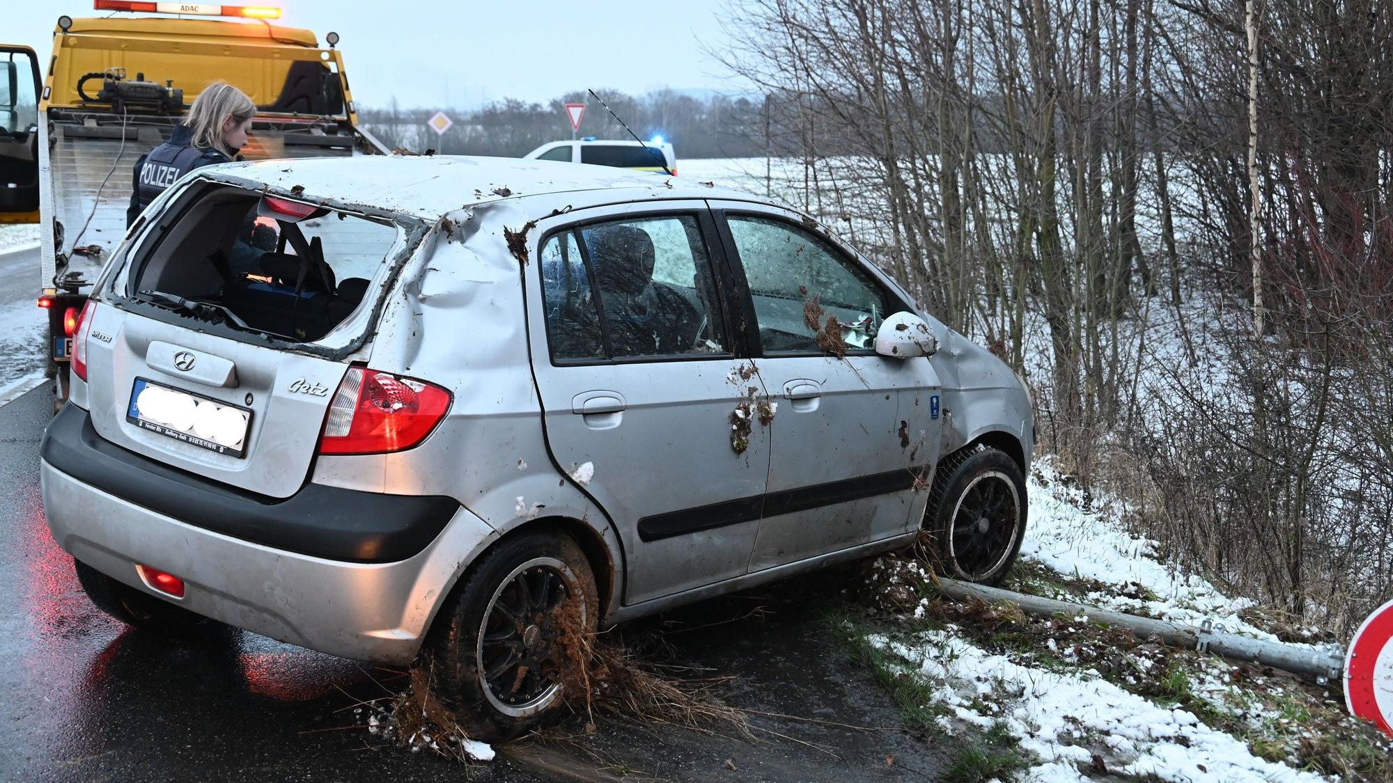 Ein beschädigtes silbernes Auto steht am Straßenrand einer Landstraße. Ein Straßenschild liegt umgekippt unter dem Vorderreifen.
