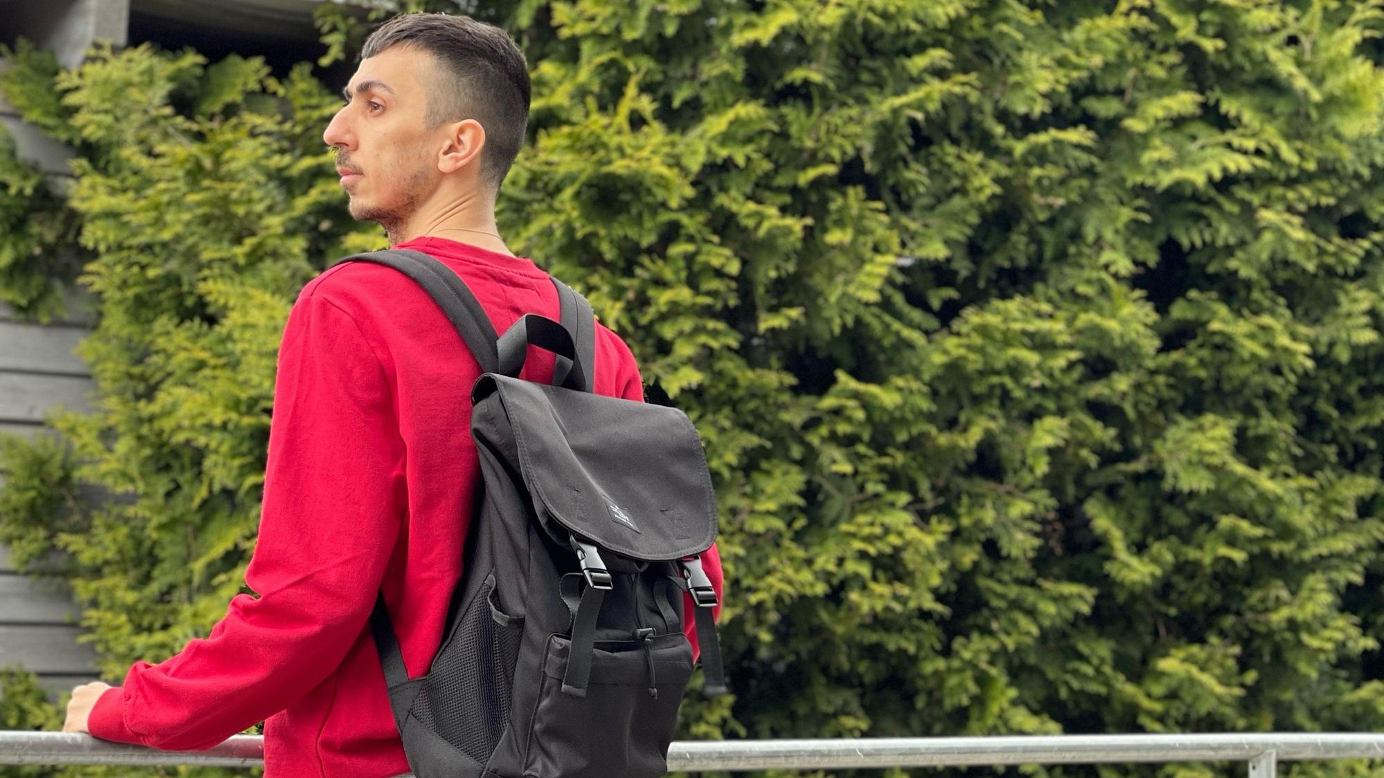 Der 33-jährige Syrer Hany steht auf einem Balkon. Er trägt einen roten Pulli und einen schwarzen Rucksack. Im Hintergrund sind Bäume zu sehen.