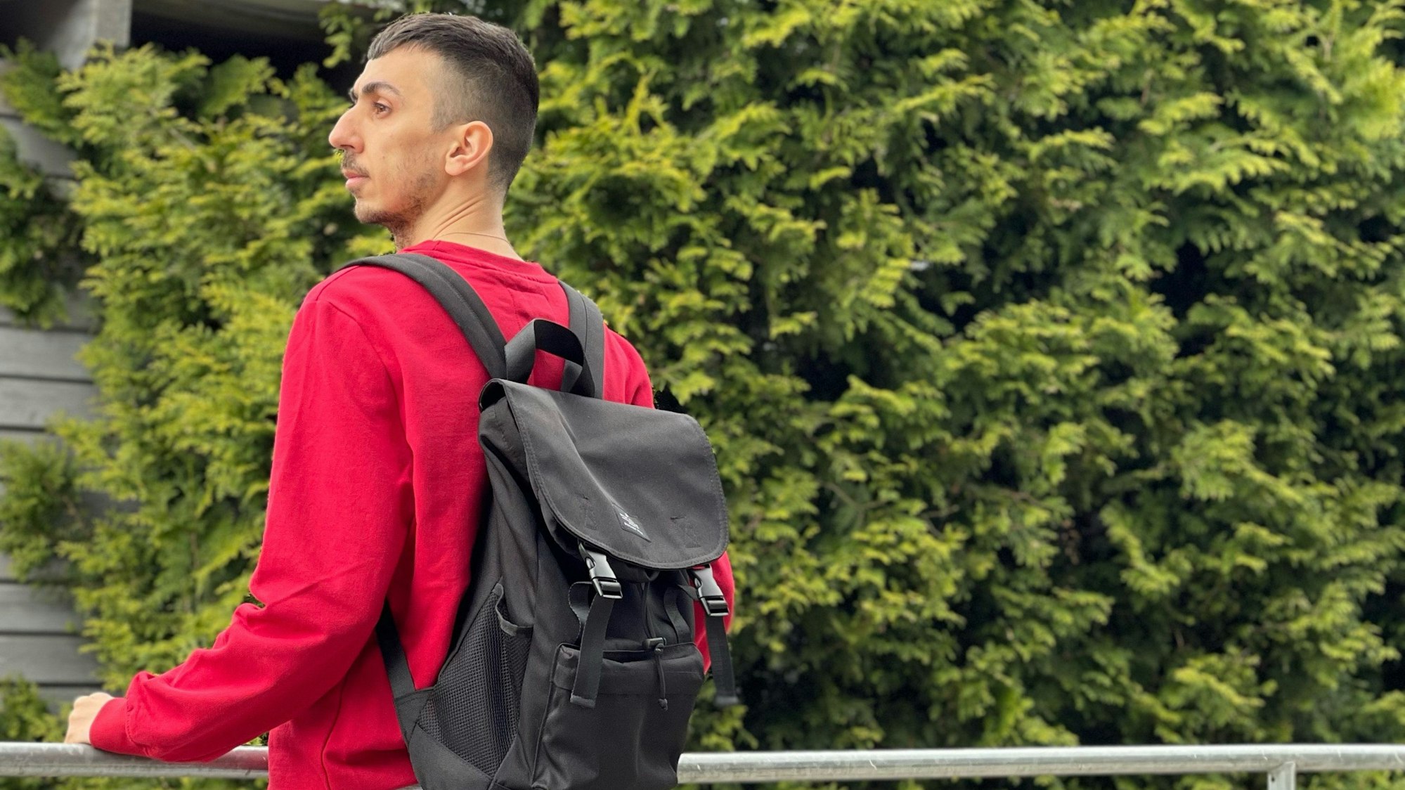 Der 33-jährige Syrer Hany steht auf einem Balkon. Er trägt einen roten Pulli und einen schwarzen Rucksack. Im Hintergrund sind Bäume zu sehen.