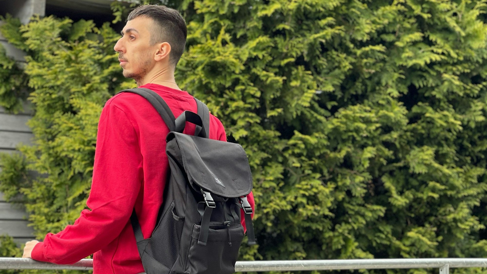 Der 33-jährige Syrer Hany steht auf einem Balkon. Er trägt einen roten Pulli und einen schwarzen Rucksack. Im Hintergrund sind Bäume zu sehen.