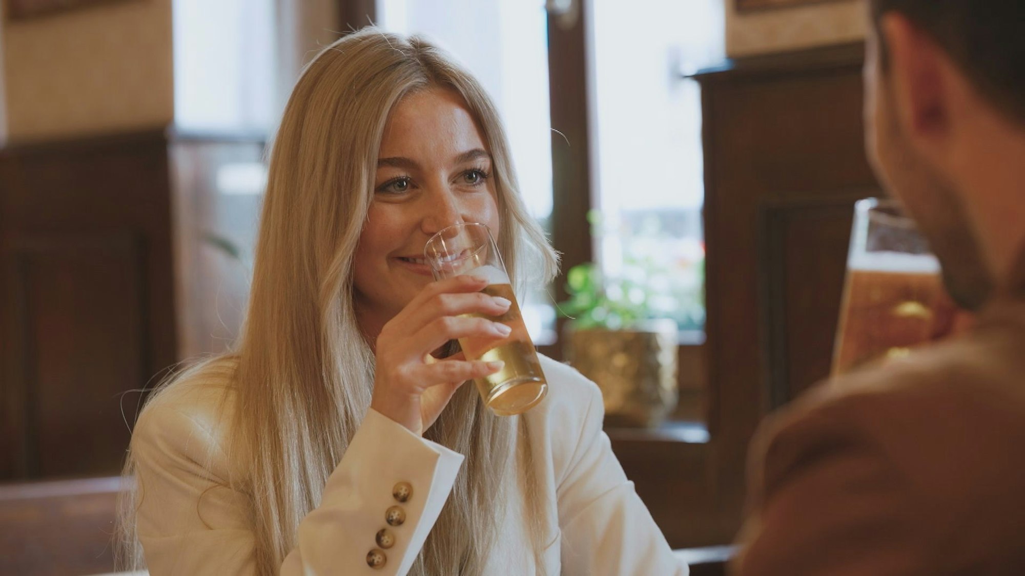 Eine Frau mit blonden Haaren hat ein Kölsch in der Hand und lacht ihr Gegenüber an.