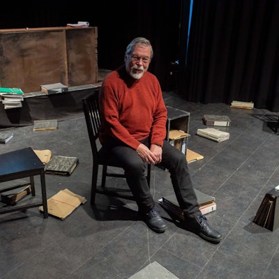 Raimund Binder sitzt auf einem Stuhl auf einer Bühne. Um ihn herum im Bühnenbild herrscht Chaos. Überall liegen Kladden und Ordner.