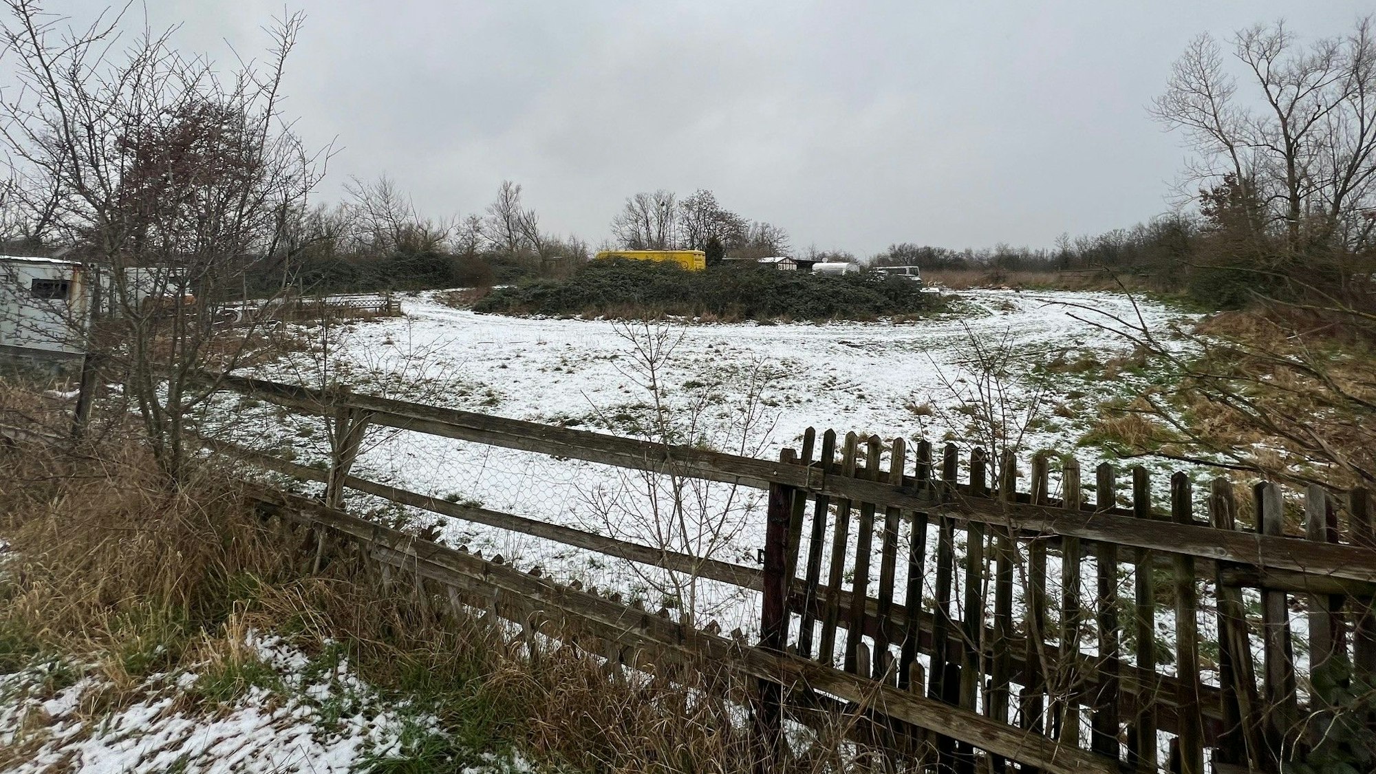 Auf einer verschneiten, von einem Holzzaun umgebenen Wiese in Rath-Heumar stehen Baracken und ausrangierte Fahrzeuge.