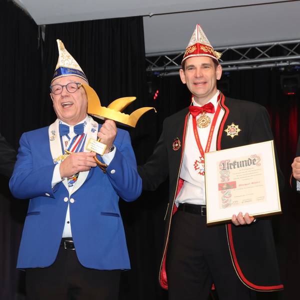 Drei Karnevalisten stehen beisammen. Der Mann in der Mitte hält eine Auszeichnung in Form einer goldenen Narrenkappe in den Händen.
